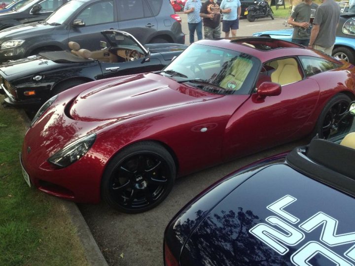 South Bucks Car meet Tuesday 13th August - Page 1 - Thames Valley & Surrey - PistonHeads - The image captures a convoy of sports cars parked on a road. The cars are lined up, occupying both sides of the road. One car, prominently in the center of the image, stands out with its red and black color scheme. People can be seen standing around the cars, possibly admiring or discussing them. In the backdrop, green grass provides a contrasting environment to the urban setting of the road.