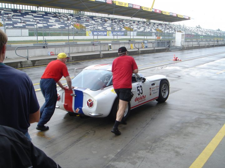 Early TVR Pictures - Page 25 - Classics - PistonHeads - This image captures a moment on a race track where two individuals are inspecting a striking white and blue convertible sports car. The car, adorned with orange racing stripes and the number 53, is parked on the wet track. A third person, amidst a backdrop of empty seats, appears to be assisting with the inspection process. The scene is set against a backdrop of a gray, cloudy sky, adding a sense of anticipation to the setting.