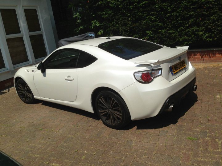 RE: Toyota GT86 vs Subaru BRZ: Blood Brothers - Page 2 - General Gassing - PistonHeads - The image shows a white car parked on a driveway. The car is a modern coupe with a sleek design and a black roof. It has a spoiler on the back and a single round taillight on the left side. The wheels have black rims, contrasting with the car's white color. Behind the car, there's a building with a white exterior, and a covered outdoor seating area can be seen. The driveway has a brick pattern, and there is bright sunlight casting shadows on the ground.