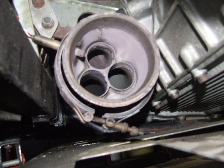 Pistonheads Lambda Sensor - The image shows a close-up view of an engine or mechanical part with a large cylindrical component at the top. This central part appears to be composed of multiple rows of elongated holes or perforations. The component is affixed to other metallic components by a series of bolts. The overall image is taken in a setting with industrial machinery in the background, suggesting this could be a workshop or a maintenance area for vehicles. The focus of the photograph is sharply on the central component, with the edges and background details appearing blurred due to the shallow depth of field.