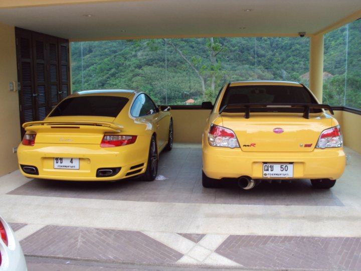 Problems Ford Pistonheads Focus - The image shows an outdoor parking area in front of a covered space. There are two yellow sports cars parked side by side, positioned as if they are facing each other. Both vehicles are branded, indicating they could belong to car enthusiasts. The setting suggests a residential or possibly a resort area, given the presence of a staircase and a partial view of greenery. The weather appears to be sunny.