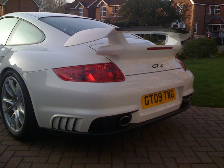 show off your porsche - Page 11 - Porsche General - PistonHeads - The image depicts a parked white sports car, which appears to be a Porsche given its recognizable design. The car's rear witch tail light design is visible, and the trunk area is adorned with a wing and the GT2 model designation. In the background, there is a well-maintained green lawn, a pathway, and nearby residential structures. The photo captures the car in daylight, and the composition emphasizes the rear of the vehicle, evoking its sporty appeal.