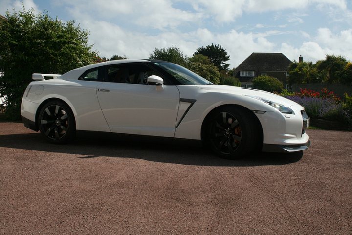 A car parked in a parking lot next to a car - Pistonheads - The image captures a scene of a white Nissan GT-R sports car parked in a driveway, facing to the left. The car has a sleek design with black rims and a visible rear spoiler. The rear of the car prominently displays a Nissan badge and dual exhaust pipes. The sky is clear and blue in the background, and there are bushes and flowers visible in the vicinity of the car. The setting appears to be a residential area during daylight hours.