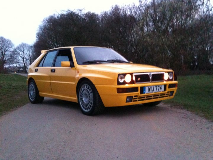 Pistonheads Endulge Seconds - In the quiet of the evening, a vintage yellow coupe caroons along a serene road. Partially obscured by the car's diagonal setup, a single, vibrant tree stands sentinel next to the roadway. The scene is encapsulated by the dimly lit road stretching out in front of the car.