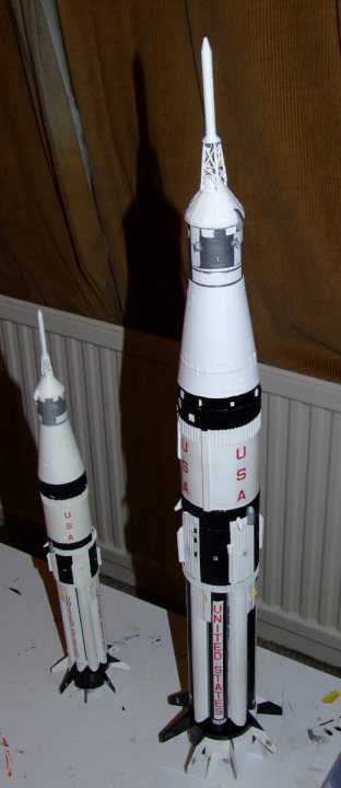 Apollo Saturn Pistonheads - In the image, there are two model rockets glued together. The larger model rocket occupies the upper two-thirds of the frame, standing on a table. The smaller model rocket is positioned towards the lower third of the image, displayed on a different surface. Below the larger rocket, a piece of text is visible, possibly indicating the name of the rocket or the mission it simulates.