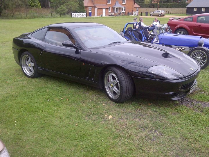 Pistonheads - In the image, a sleek black sports car is parked on a lush green lawn. The car, with its shiny exterior reflects the clear sky above. It's situated in a field with other vehicles, including a blue and a red one, partially visible behind it. The scene is vibrant and outdoorsy, with the car as the main focus against the expansive green backdrop.