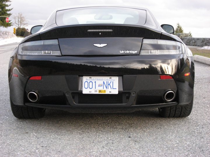 My PDC - Page 1 - Aston Martin - PistonHeads - In the heart of a snow-laden landscape, under a gray sky, a sleek black LeGrand sports car rests on a quiet road. The car's license plate, reading "001 KNL", stands out against its glossy coating, subtly marking its identity. The emblem of a silver bird graces the trunk, hinting at a touch of luxury and power that lies within the car. The scene is serene, with the car's design and presence adding a touch of life to the otherwise still and white winter landscape.