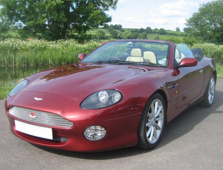 Being Bold..... - Page 2 - Aston Martin - PistonHeads - The image shows a red convertible sports car parked on what appears to be an asphalt surface. The car's top is down, revealing a sleek interior. The vehicle features a front grille with a logo that resembles a stylized wing, indicative of a model by Aston Martin. It has a glossy finish, and the design suggests a modern and high-performance vehicle. The background includes a grassy area and trees, providing a natural setting for the car.