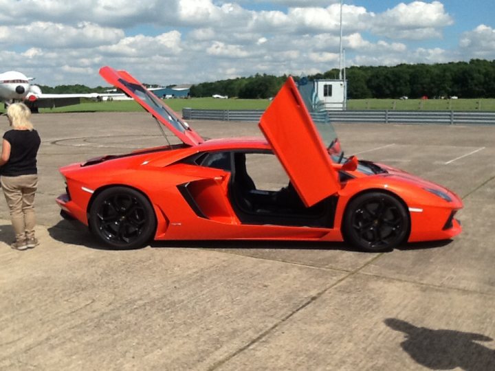 Aventador at Bruntingthorpe - Page 1 - Diablo/Murcielago/Aventador - PistonHeads - This image captures a vibrant scene at an airport tarmac. Dominating the frame is a striking red Lamborghini sports car, parked on the expansive concrete runway. The car's hood is artfully open, revealing the engine within, while its sleek black wheels and red tail lights stand out against the vehicle's color.

In the background, several airplanes are parked on the tarmac, their metallic bodies gleaming under the daylight. A few seemingly unrelated objects, including a bottle and a suitcase, are scattered in the vicinity, hinting at the activity on the tarmac.

A person, wearing a casual black T-shirt and gray trousers, stands beside the car, their attention seemingly drawn towards the plane in the distance. Their presence adds a touch of human element to the otherwise mechanical landscape of the tarmac.

Above everything, a blue sky stretches out, dotted with fluffy white clouds. This natural elements provide a serene contrast to the technological marvels displayed on the ground.