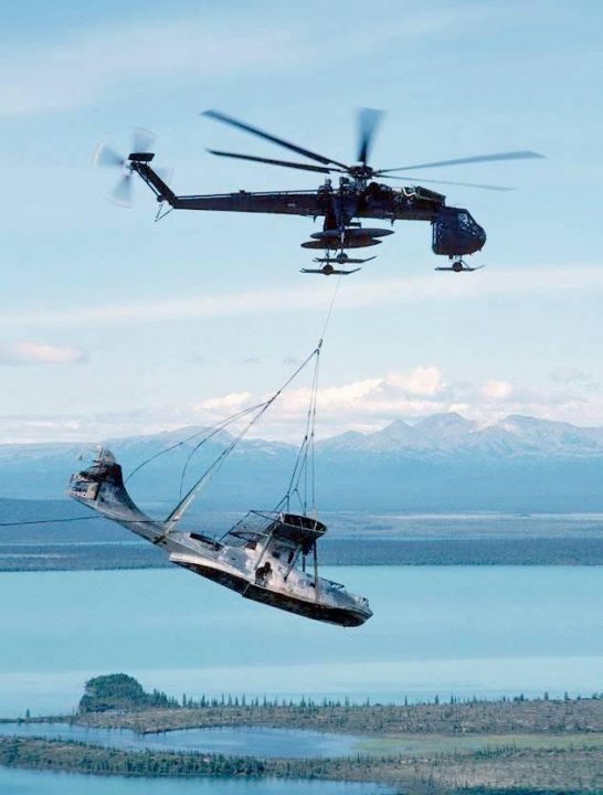 Post amazingly cool pictures of aircraft (Volume 2) - Page 415 - Boats, Planes & Trains - PistonHeads - The image presents a striking scene of a military helicopter in flight, carrying an assault boat in its cargo bay. Below the helicopter, a crane is positioned to unload the boat into the water below. The backdrop of the scene is a serene landscape featuring calm waters and distant mountains under a clear blue sky. This image captures the precision and logistics involved in military operations.