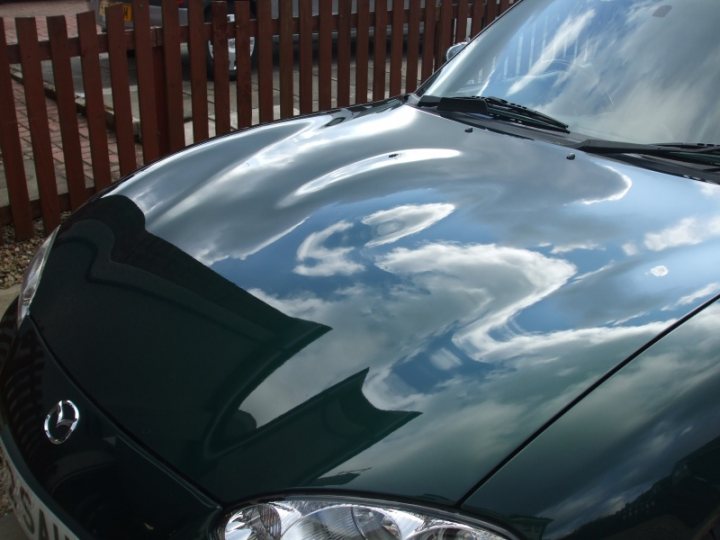 Pistonheads Shiney - The image features a shinny car parked in front of a wooden fence. The reflection of the sky on the car's surface is striking, with the pattern of clouds mirrored on the polished surface of the hood. The scene creates a stark contrast between the stillness of the vehicle and the dynamic, organic shapes of the cloud reflections. The setting is tranquil, with no people in sight, emphasizing the car's reflective surface as the main subject of the image.