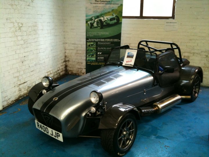 Pistonheads - The image showcases a futuristic-looking vehicle, which appears to be an early model of a Lotus Elise. Its distinctive design features a sleek body with a vibrant yellow and black stripe running down the center. It's equipped with classic black wheels and a prominent, round headlight. The vehicle is displayed in a white room with blue walls, positioned next to a green information board. The Elise is likely an item for sale, given the "SOLD" sign on the windshield.