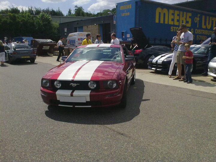 Bbq Atlantic July Sat Pistonheads Sports - The image shows a vibrant red sports car with a distinct white stripe on the hood, parked on a tarmac. There's a group of people standing nearby, engaged in various activities. In the background, on the right side, there are several other cars parked inside a fenced lot. A blue truck with the words "MEEKS OF LUTO" is also visible in the background. The weather appears to be sunny, and the setting seems to be an outdoor lot or parking area.