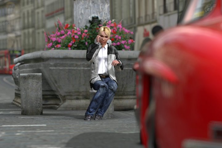 Guess Pistonheads - The image showcases a city scene with a focus on a woman sitting on a bench. She is holding a phone to her ear, seemingly engrossed in a conversation. Behind her, a beautiful flower arrangement adds a touch of nature to the urban setting. The bench is situated next to a road, with a red car partially visible, indicating that she might be waiting for it to pass before she continues her journey. The image captures a moment of urban life and communication.