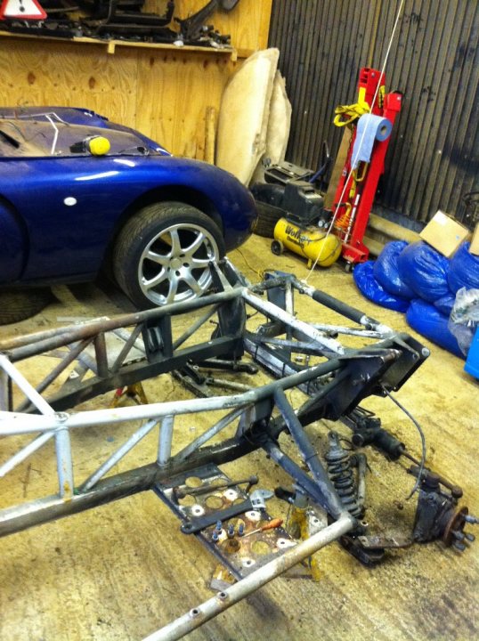 Dissassembly of Tuscan is complete.....what now ???? - Page 1 - General TVR Stuff & Gossip - PistonHeads - The image displays a concrete area containing various parts and tools designated for use in a garage or workshop. In the background, there is a blue sports car propped up on a jack, supported by a piece of custom-built metal frame. Scattered around the car, there are wheel rims, mechanical parts, and some junk. On the floor, there's a yellow air compressor and a red pressure washer, suggesting this could be a workspace for maintenance or repairs. At the bottom of the photo, a metal frame appears to be the chassis of a race car, indicating the area could be a workspace for repairs or customization.