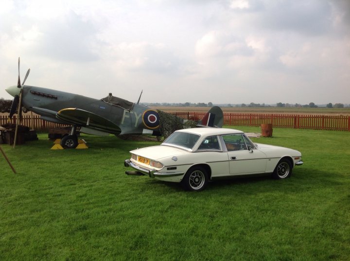 Triumphs and Spitfires - Page 1 - Triumph - PistonHeads - The image showcases a vintage setting where two historical vehicles, a car and a plane, are on display. The car, front and center in the frame, is a classic sports car with a distinctive circular emblem on its grill. Just behind the car, parked diagonally and slightly elevated, is a green and gray fighter plane featuring a large glass window in the cockpit area and a smaller one behind it. The vehicles are set against a backdrop of a grassy field with a fence running along the horizon. There is a ficus or similar tree in the lower left corner of the image, adding a touch of greenery to the scene. The contrast in color between the vehicles and their surroundings, along with their distinct shapes, make them the undeniable focal point of the image.