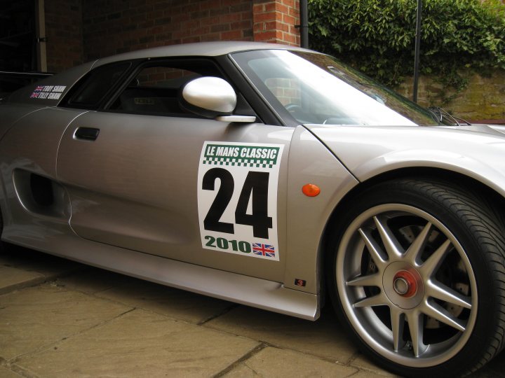 Pistonheads Stickered - The image shows a silver sports car parked on a brick-paved street. The car is painted a sleek silver and features a numerous brand identification. It is parked next to a wall, and a sticker on the car displays "Le Mans Classic 24 2010" with a British flag, signifying its participation in an event at the famous racing circuit. The car's wheels and suspension are detailed in the photo, highlighting its aesthetic appeal and racing capabilities.