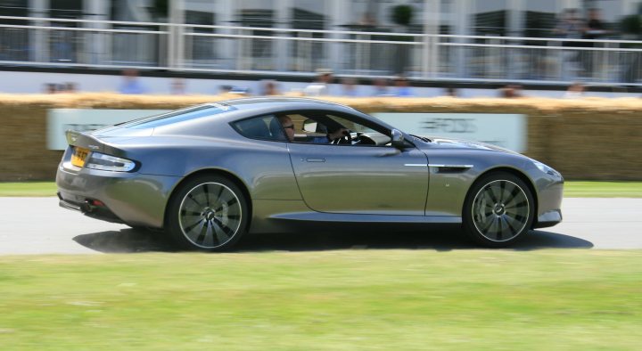 Goodwood moving motor show photos - Page 1 - Goodwood Events - PistonHeads - The image shows a dark grey sports car in motion on what appears to be a race track. Seated in the car is a man, presumably the driver. The vehicle is shiny, indicating either a recent clean or its good condition. The photo is clear, capturing the car as the central focus against a blurred background of a race track and spectators.