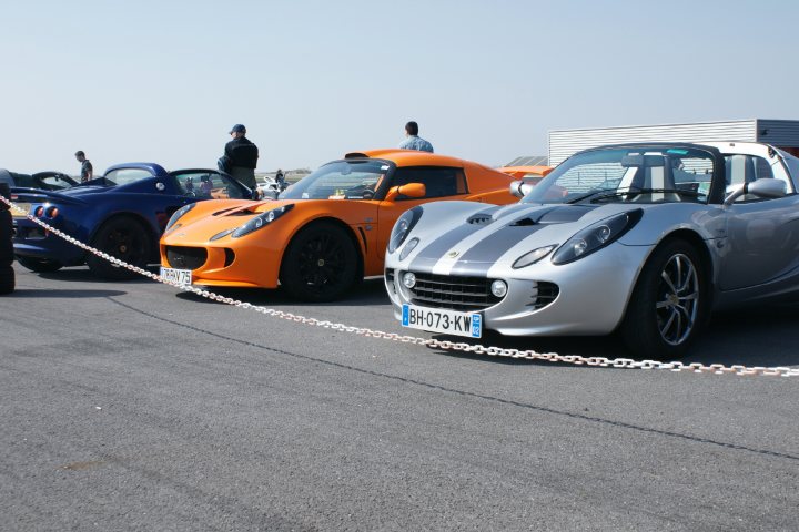 Chrome Orange Exige pictures please !! - Page 1 - Elise/Exige/Europa/340R - PistonHeads - The image depicts a vibrant scene at what appears to be a track day or a similar motorsport event. Four stylish, modern sports cars are parked in a line, each with a distinct color: blue, black, orange, and white. They are mounted on slick tires, which are often used at racetracks for better grip and performance. 

In the foreground, a chain restricts access to the cars, indicating that this might be a limited or private event. There are people in the background, appreciating the gathering of these beautiful machines. Their presence suggests that this event is open to the public, and enthusiasts have come to admire, photograph, or participate in the activities. The setting is an outdoor track, and the clear sky indicates good weather for this outdoor event. The image captures the excitement and passion for motorsports.