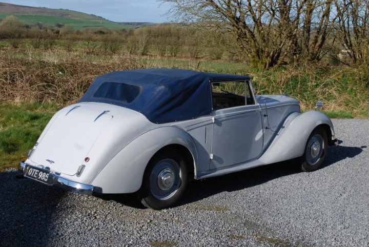 Anyone remember this car from the 1950s or 60s? - Page 1 - North West - PistonHeads - The image showcases a classic white car with a protective blue canvas top, parked on a gravel surface. The car features an antique design with rounded fenders, a vintage steering wheel, and a set of curved headlights and a trunk. The gravel surface suggests a rural or off-road setting, and there are large trees with abundant foliage in the background, which implies that the photo was taken during the spring or summer season. The lighting indicates a clear day with no visible clouds.