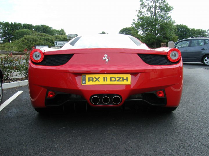 Breakfast Meeting, The Hare, Roxwell 4th June - Page 1 - Kent & Essex - PistonHeads - The image shows a red Ferrari parked on a road. The vehicle's registration number is displayed at the back as "RX II DZH". The Ferrari's brake lights are prominent, with two red lights visible on each of the back side lights. The setting appears to be during the daytime, with a partially visible gray car in the background. The scene has a clear sky in the upper left corner, implying fair weather conditions.