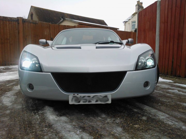 JFReturn's VX220 2.2 NA - Page 1 - VX - PistonHeads - The image shows a close-up view of the front of a white sports car parked on a driveway. The car has a sleek, aerodynamic design with a prominent grille and sharp headlights. Its hood has air vents, and there's a visible sweat print on the right side of the hood. The vehicle is positioned at an angle, slightly to the right, providing a clear view of its front and part of its right side. The image is a bit grainy, suggesting a low light or a higher ISO setting was used while taking the photo.