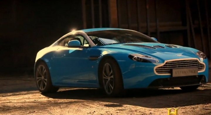 Vantage S on the telly - Head Up - Page 1 - Aston Martin - PistonHeads - The image displays a striking blue Aston Martin car, identified by its license plate. The vehicle is parked on a dirt ground, somewhat off the road. The setting appears to be outdoors during the morning or late afternoon, as suggested by the golden light lighting up the car's exterior. The backdrop is quite plain, with a blurred texture that could be a wall or a barrier, ensuring the focus remains on the vehicle.