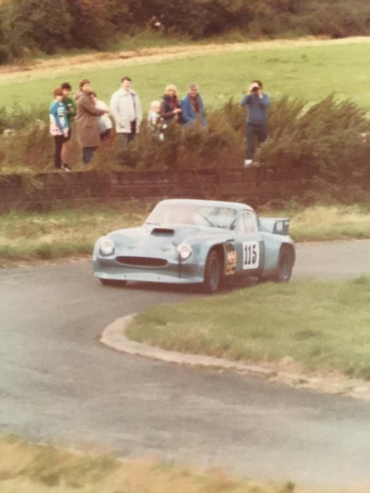 Early TVR Pictures - Page 114 - Classics - PistonHeads - In the image, a vintage sports car is in motion, rounding a right turn on a race track. The car is in the foreground, with its headlights shining brightly. A group of spectators is scattered around the track, watching the car race at high speeds. The setting is outdoors, and the spectators appear to be a mix of standing and sitting, with some holding bottles and others using cameras to capture the thrilling moment. The ground of the track is grey, and the area surrounding it is partially covered in grass and shrubs.