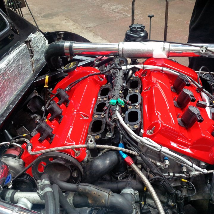 Engine Bling - Page 1 - Noble - PistonHeads - This image showcases an open, dirty, and extensive portion of the engine bay of an engine. The main focus is on a red fuel pump, which is attached to the engine via a large metal pipe, surrounded by an array of hoses, wires, and other engine components. The scene appears to be a mechanical workshop, given the absence of the engine's hood and the presence of hoses and wires that haven't been tucked away or finished. There are no visible texts or branding within the image.