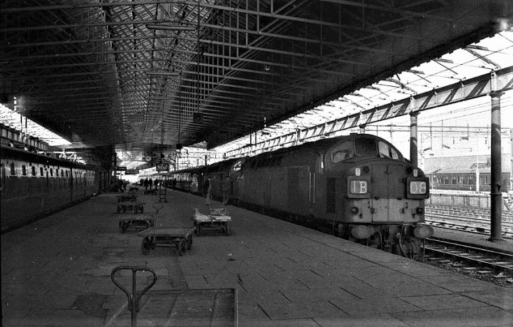 Loco sheds and other railway buildings.... - Page 4 - Boats, Planes & Trains - PistonHeads UK - The image is a black and white photograph depicting an interior train station. A large train with multiple carriages is parked at a platform, spanning the length of the station. Above the train, a metal walkway leads to additional platforms or stations not visible in this frame. On these upper platforms, smaller trains can be observed, suggesting multiple lines passing through this terminal. The station itself appears empty, with no passengers in sight, and is characterized by concrete flooring and a high ceiling.