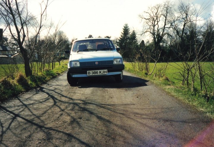 Thirty years old and still going strong :~ - Page 1 - MG - PistonHeads - In the image, a silver Volkswagen export is parked on the side of a gravel road in the suburbs, overlooking a scenic view of trees and foliage. The car features a European license plate, suggesting it has been recently imported. The road appears to be somewhat narrow, with the car taking up a significant portion of its width. The surroundings are lush with nature, indicating a peaceful and serene environment.