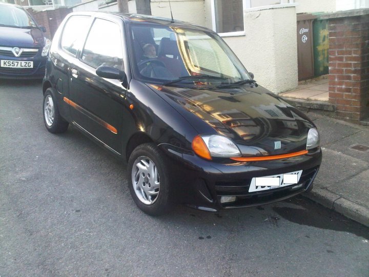 Love Pistonheads Shed - The image features a compact black car parked on the side of a street. It has a registration plate that reads "LJ57 TJZ," and there is a person partially visible inside the car. The car's design suggests it's a small hatchback model, and the context implies a suburban or city environment. The sky visible in the background is partly cloudy.