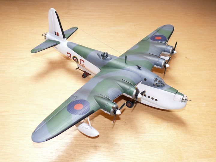 Sunderland Pistonheads Shorts - The image features a model airplane, specifically a World War II fighter plane, painted in a camouflage pattern with shades of gray, green, and white. The aircraft is equipped with two sets of black propellers, and the landing gear is indicated by white extensions. Its design includes a forward-facing gun turret and a rear fuselage gun port, both of which are colored red. The plane appears from a side angle, allowing for a clear view of its distinctive features. The model is set against a wooden surface, which serves as a backdrop for the aircraft.