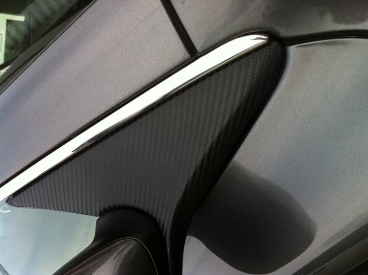 Wrapped DB9 Door Mirror Surrounds  - Page 1 - Aston Martin - PistonHeads - The image shows a close-up view of the inside of a car's door handle. The door handle is predominantly matte black with a hint of carbon fiber detailing on the bottom part. Shiny silver metal accentuates the spine of the handle. The surface texture suggests a durable and modern design, while the metallic edge has a smooth, polished appearance. The reflection of a slightly cloudy sky is visible in the window, providing context about the location and weather during the photograph.