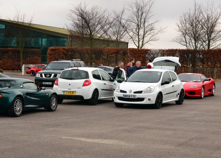 Robert Barrie Track day 24th Feb - photos - Page 1 - Goodwood Events - PistonHeads - The image shows a parking lot filled with various cars. The cars range in size and color, making a diverse display. Some of the vehicles are public transit buses and sedans, while others are more unusual, including coupes and sports cars. The lot appears to be relatively open with hedges in the background. There are a few people scattered around the lot, though they are mostly outside of the frame, and not the main focus of the image.