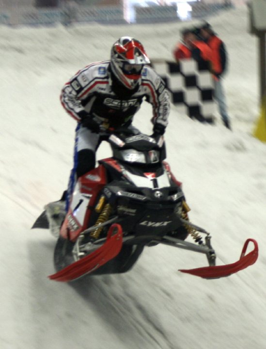 The image features a thrilling scene of a person competing in a snowmobile race. The snowmobile, with prominent branding, is in full motion on a snow-covered track, kicking up a cloud of snow. The driver, clad in a snowsuit and goggles, leans forward with a determined expression, demonstrating skill and focus as they navigate around a bend. In the background, the blurred presence of other competitors and track officials indicates an intense competition in progress. The atmosphere electrifies the viewer, perfectly capturing the adrenaline-fueled energy of the sport.