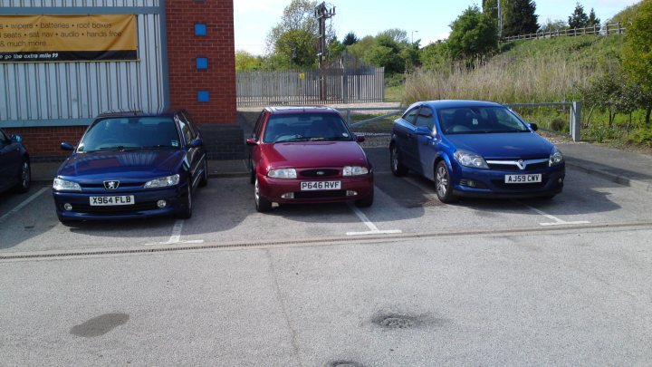 Pistonheads - The image shows a parking lot with three cars parked parallel to each other. On the left is a dark blue Honda car, next to a red compact car, and on the right is a metallic blue vehicle, which appears to be a newer model. The cars are positioned in front of a sign and a fence. The sign has text on it, but it's not legible in the image. The setting suggests it could be daytime with overcast or cloudy weather. The photo seems to have been taken from across the parking lot, giving a clear view of the vehicles and the immediate surroundings.