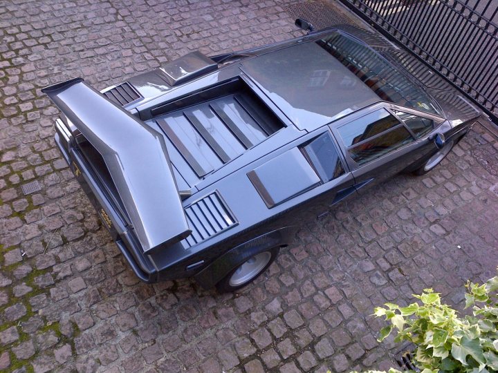 Countach  - Page 54 - Lamborghini Classics - PistonHeads - The image shows a dark-colored storage container covering the cargo area of an old, possibly sixties, car. This decapsulated vehicle has been modified to include an open-air cargo space, which is enclosed by a weathered storage container. The car itself is black, and the storage container has a prominent, upturned rear section. The vehicle is parked on a cobblestone surface, suggesting the image may have been taken in an urban or historic setting. In the background, a portion of a black gate is visible.