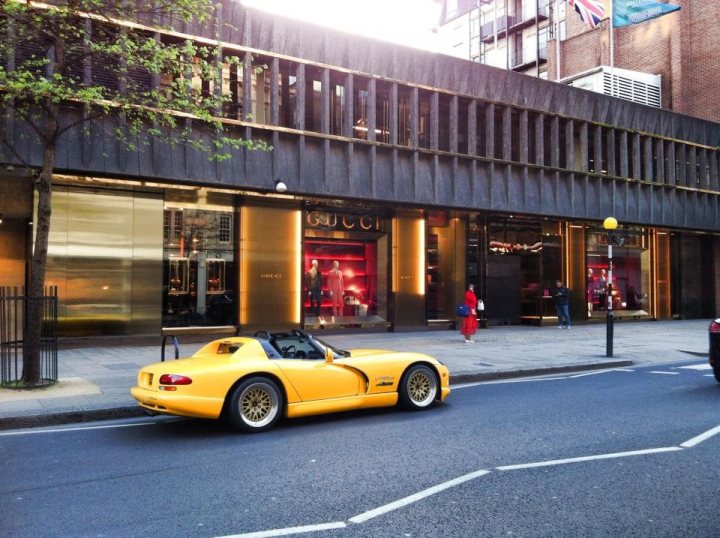 Ready for the summer - Page 1 - Vipers - PistonHeads - The image captures a scene on a city street. A bright yellow sports car is parked on the side of the street, adjacent to a building. The building has a distinctive black metal facade, and there is also a tree visible in the surroundings. There are a few pedestrians walking on the sidewalk near the building, adding a sense of activity to the scene. Additionally, there is another, smaller car parked at an angle in front. The background features a large open space with a hint of green on one side, suggesting the street is situated in an urban environment.