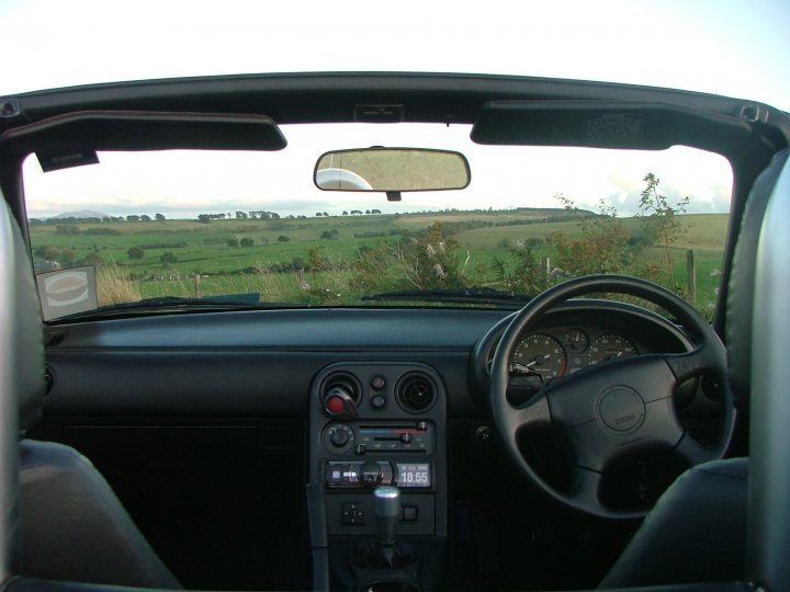 Show us your MX-5 .  56k run away - Page 35 - Mazda MX5/Eunos/Miata - PistonHeads - The image provides a view from the driver's seat of a convertible car, with the backdrop featuring a peaceful countryside. The interior of the car reveals a clean and well-maintained environment. The car features a convertible soft-top top, which is closed, and the seats are black and in good condition. On the dashboard, there is a display of sliders, dials, and indicators, indicating the settings or dashboard information. The ambiance of the image suggests a calm and serene driving experience, possibly on a leisurely road trip through the countryside.