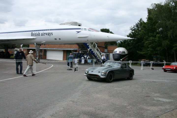 Early TVR Pictures - Page 126 - Classics - PistonHeads - The image captures a unique scene of a green Connaught car on a gray paved lot. The car is positioned imposingly in front of a British Airways jetliner. The jetliner is a white and gray paneled Concorde, with its nose tilted upward as if ready for flight. The jetliner is a marvel of engineering, parked in front of a concrete building. In the background, two individuals can be seen crossing the lot, adding a sense of scale to the image. The overall scene is a blend of modern technology and public access, with the Concorde serving as a symbol of both eras.