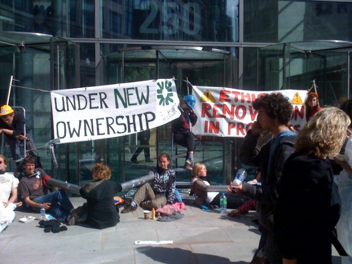 Climate Rbs Nobbers Pistonheads Update - The image shows a group of people congregating on a sidewalk with clear glass doors in front of them. The people are holding up white signs with black handwriting decrying the prospect of "under new ownership". One person near the edge of the frame is holding a water bottle, and another person closer to the camera appears to be holding a paper or card. There is no visible text on the signs that would reveal any specifics about the ownership or the event. The scene suggests a gathering or protest of some kind, although the exact nature of the event or movement is not clear from the image itself.