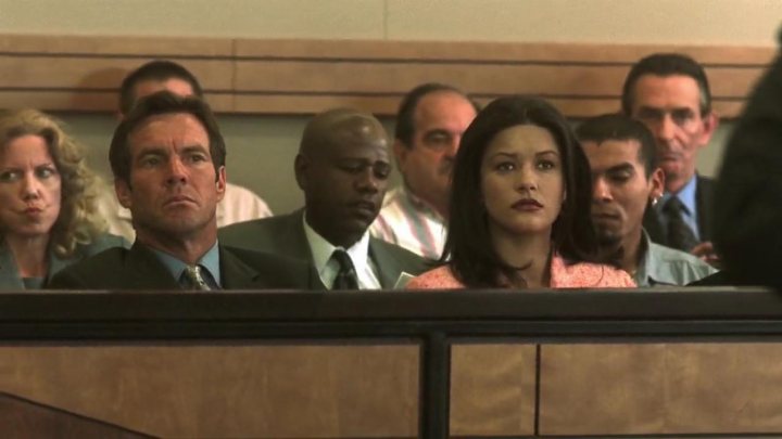 The image shows a group of people in a courtroom setting, looking attentively ahead. One individual in the front of the group is prominently staring ahead with a serious expression, while the rest of the group is slightly turned to their right. The group consists of both men and women, and some of the men are dressed in suits. The backdrop suggests a formal environment, likely a law or legal proceeding.