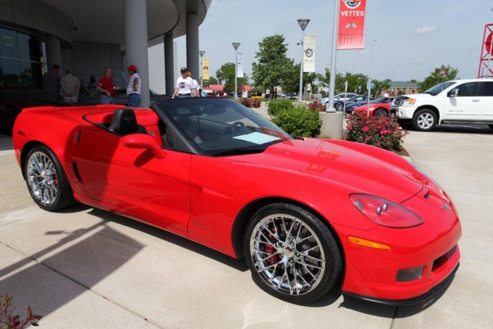 Corvette challenge what would you change? - Page 1 - Corvettes - PistonHeads - The image serves as a promotional photo for a Corvette, a notable sports car made by Chevrolet. The vehicle is parked on a sidewalk next to a dealership, which features a red banner and a car wash labeled "Vettes." The car is positioned on the right side of the photo, and a person in a white shirt is nearby, possibly a staff member or passerby. The dealership is adjacent to a parking lot where various other cars are parked, reflecting the vibrant atmosphere of a busy automotive event.