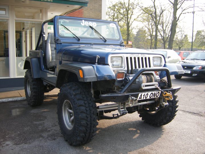 Pistonheads - The image shows a striking blue Jeep parked in an open lot. The vehicle is fairly rugged looking, featuring oversized off-road tires on a high lift kit, which adds to its robust appearance. At the back of the Jeep, there's a spare tire mounted to a tow hitch. The vehicle's windshield is not fully visible, and we can also see license plates on the front and back indicating it's been through customs; "A10 BYO" is visible. The Jeep is equipped for off-road conditions, suggesting it might be utilized for outdoor activities or exploration.