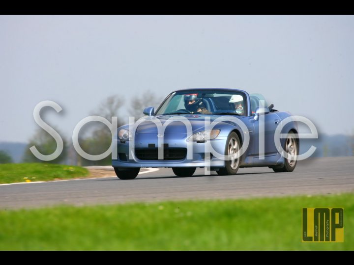 Nurburgring Pistonheads Light Stand Touchpaper Blue - This image depicts a blue sports car driving on a track or road. The car is viewed from the side, with the driver's side open to the elements. The driver is visible and appears to be wearing a dark helmet. The road is race-track smooth, and the weather is sunny with a clear sky. The term "SAMPLE" is superimposed in bold, white letters across the image in a way that suggests this is a placeholder or an example of how an advertisement might appear with the car as the featured product.