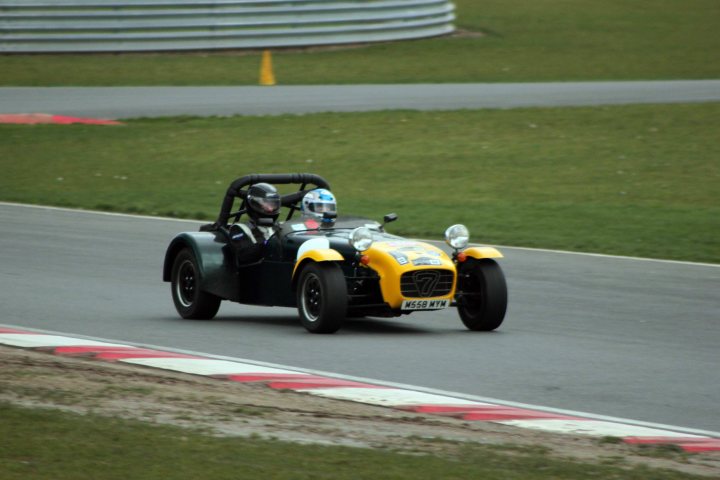 Track day at Snetterton Friday 15th February - Page 1 - East Anglia - PistonHeads - In the image, a black futuristic car is the main focus, zooming down a track. The car is sleek with copper and black accents, giving it a striking appearance. There are two people inside the car, one of whom is the driver. They are wearing helmets, indicating they are taking safety precautions. The background is a blur of greenery, suggesting high speed, and a section of metal fencing, indicating the boundaries of the race track.