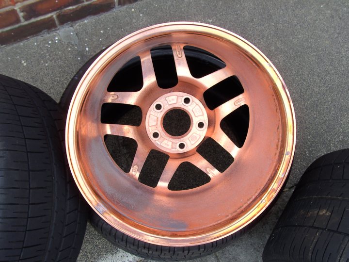 Wheel Pistonheads Chrome Refurb - The image features a single car tire placed on a paved surface, likely for a vehicle. The tire appears to be made of rubber and is covered with a metallic bronze-colored wheel rim. The wheel rim has a ten-spoke design, and the surface of the rim looks to be a brushed metal finish. The tread pattern of the tire is visible and seems to be standard for most tires. The backdrop of the image is nondescript, emphasizing the tire and wheel rim as the main subjects.