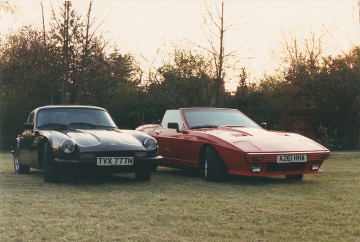 Potentially daft question - Page 1 - Classics - PistonHeads - The image captures a serene outdoor scene featuring two classic sports cars. A black car, prominently positioned in the foreground, exudes a sense of classic elegance on the grassy area with trees in the background. This sleek sports car is detailed in its color and adorns a license plate reading "TXX 77N". In contrast, the red car, parked slightly behind the black one, appears to be in a different orientation, adding depth to the scene. The presence of both cars side by side showcases a gathering of enthusiasts or perhaps a vintage car show. The image likely evokes a sense of nostalgia and admiration for these classic vehicles.