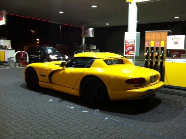 Next Ace Cafe meet Monday 9th September - Page 1 - Vipers - PistonHeads - The image depicts a night scene at a gas station. A striking yellow sports car is parked at the gas pumps, its inclusion is the main focus of the image. Other vehicles are also present, including a black sedan with a bright set of headlights illuminating the area. The station itself is illuminated by service station lights, and both gas pumps visible are branded with Shell logos.